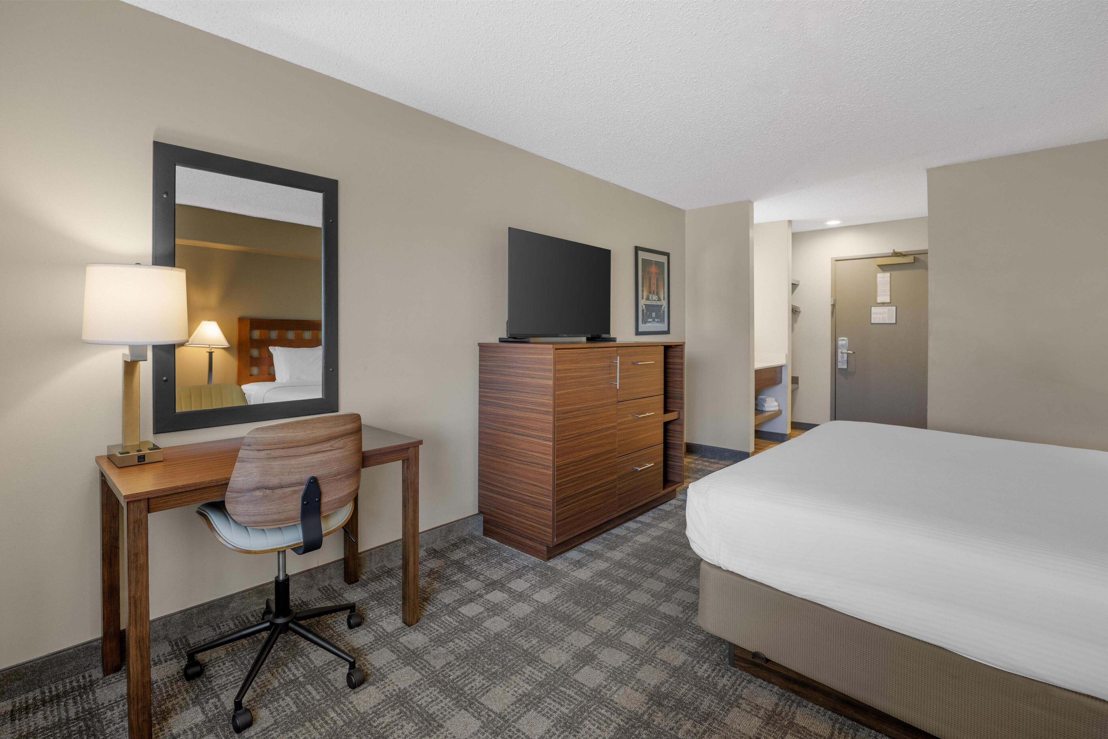 A hotel room with a bed, a desk with a chair, a dresser, a television, and a door.