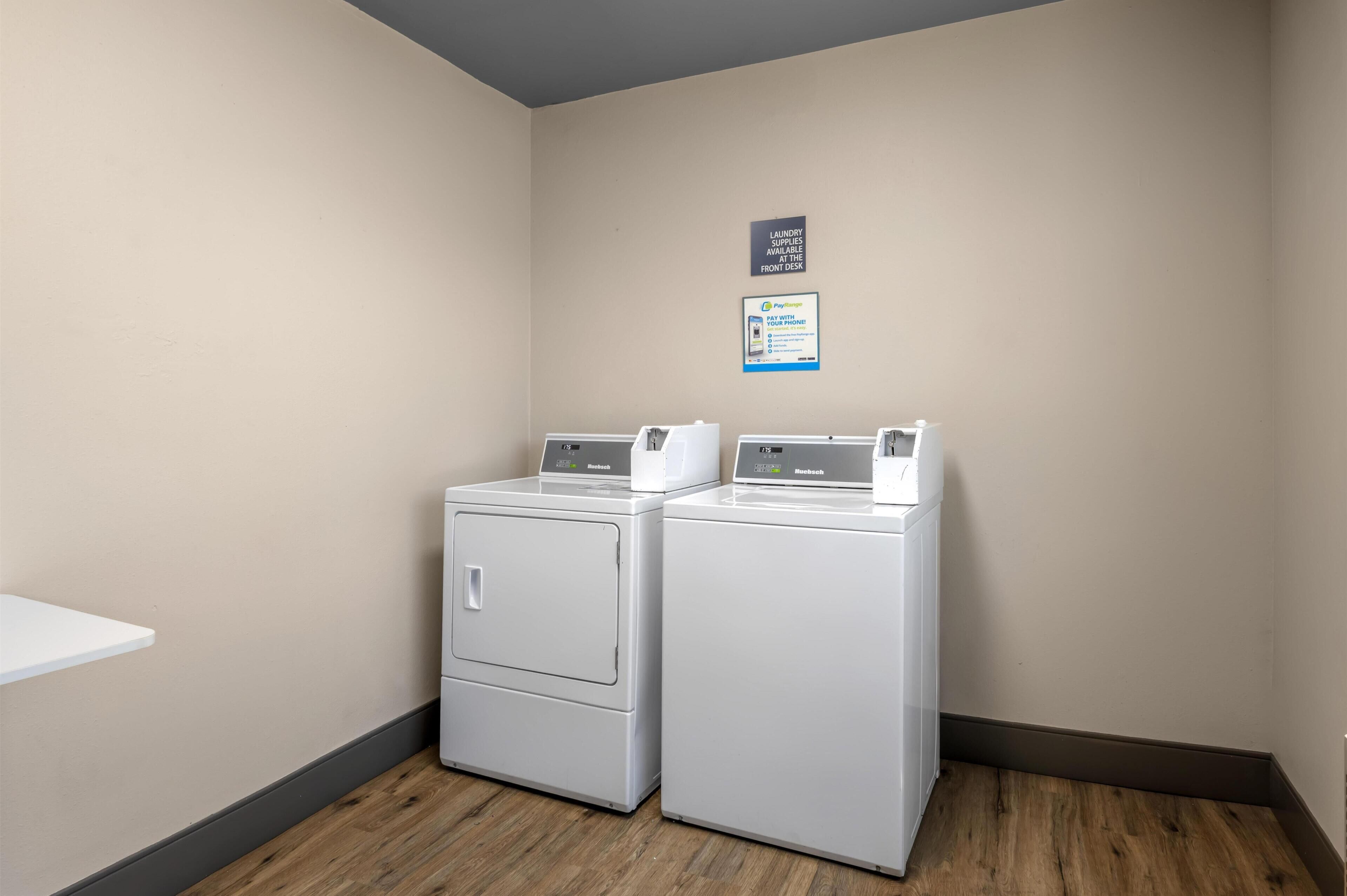 A laundry room with two stacked front-loading washers and dryers, a sign indicating laundry supplies at the front desk, and a PayRange advertisement.