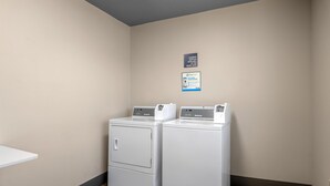 Une buanderie équipée de deux machines à laver et deux sèche-linge à chargement frontal superposés, un panneau indiquant où trouver le nécessaire pour la lessive à la réception, et une publicité pour PayRange.