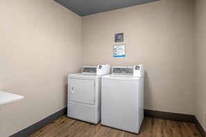 A laundry room with two stacked front-loading washers and dryers, a sign indicating laundry supplies at the front desk, and a PayRange advertisement.