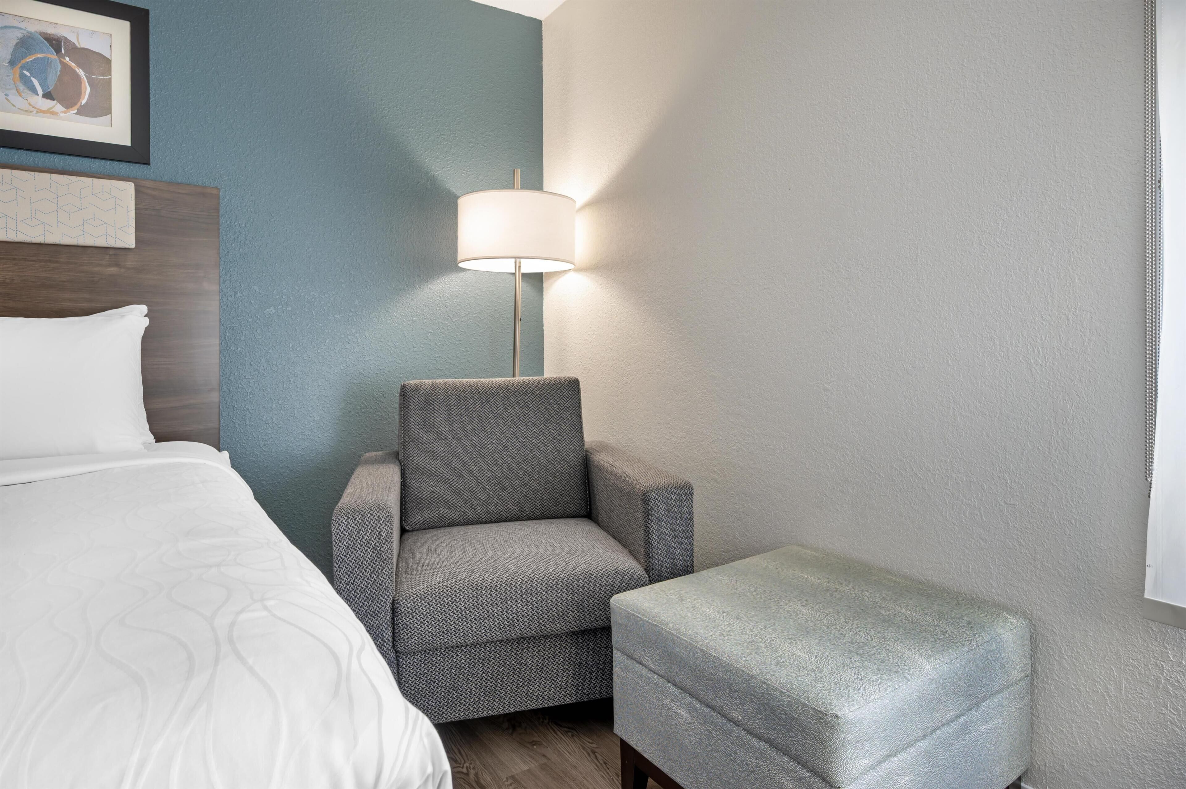 A hotel room with a bed, a grey armchair, a small ottoman, a floor lamp, and a wall with a framed picture.