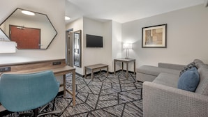 Habitación de hotel con escritorio, una silla azul, un sofá gris, una mesita, lámpara, televisor, cuadro enmarco y alfombra estampada.