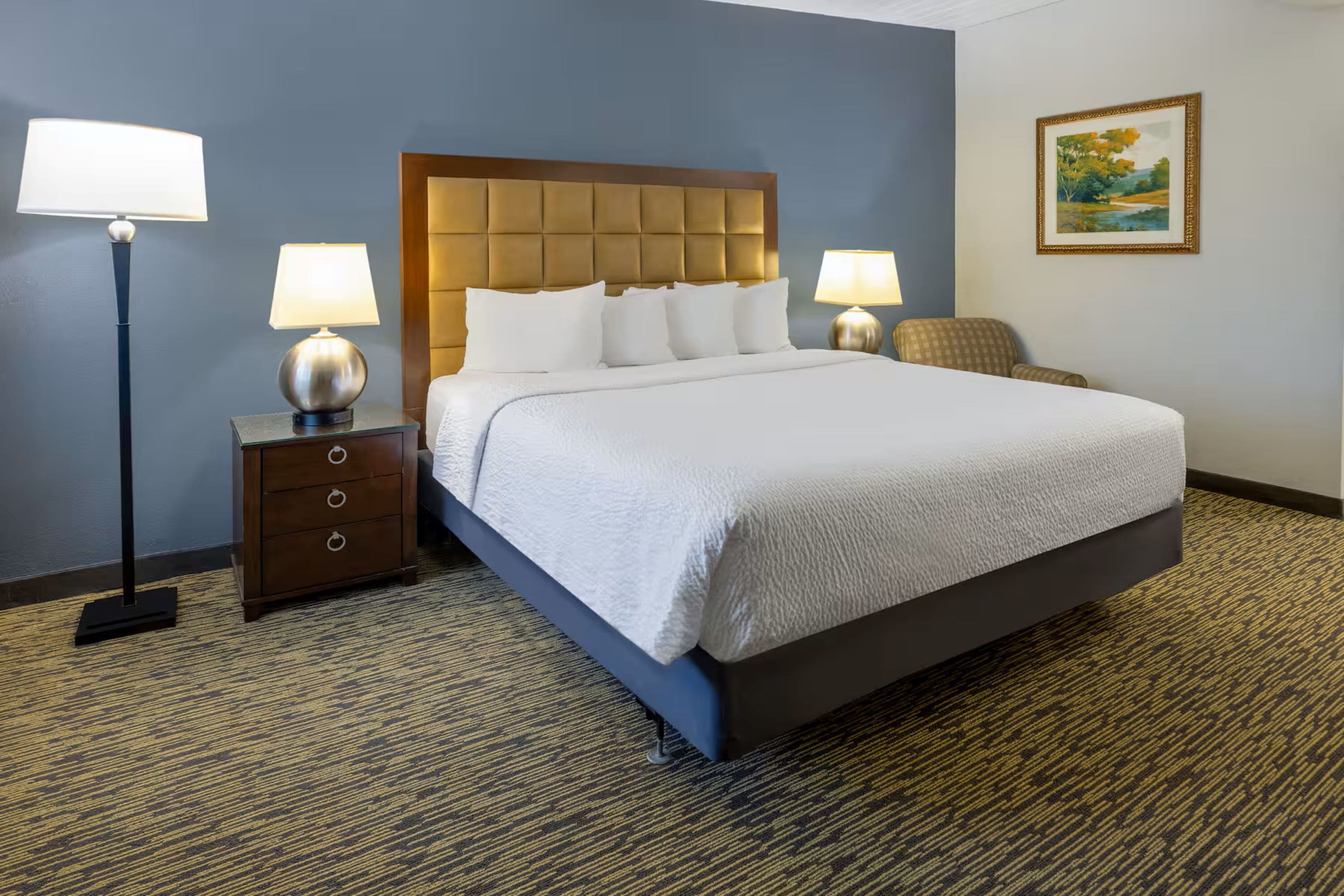 Quarto de hotel com uma cama grande, duas mesas de cabeceira com luminárias, uma cabeceira de madeira, uma cadeira e um quadro na parede.