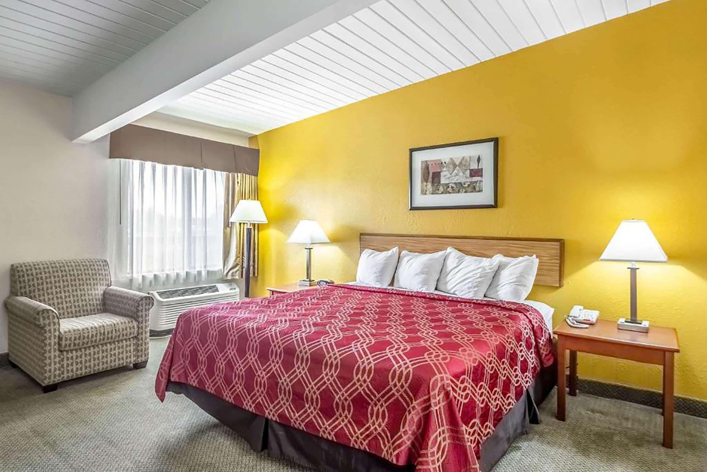 Quarto de hotel com cama, uma cadeira, mesa de cabeceira com abajur, janela com cortinas e um quadro na parede.