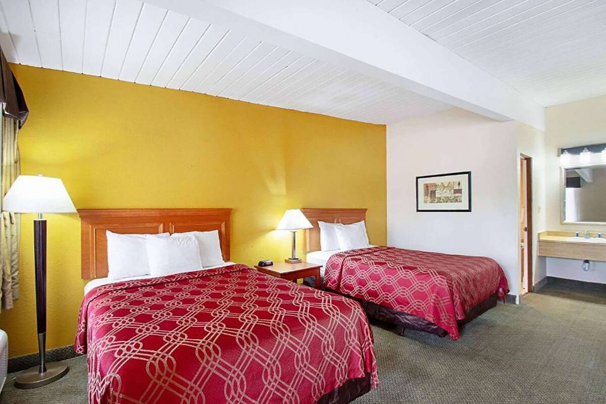 A hotel room with two beds, a bedside table with lamps, a bathroom with a sink and mirror, and a framed picture on the wall.