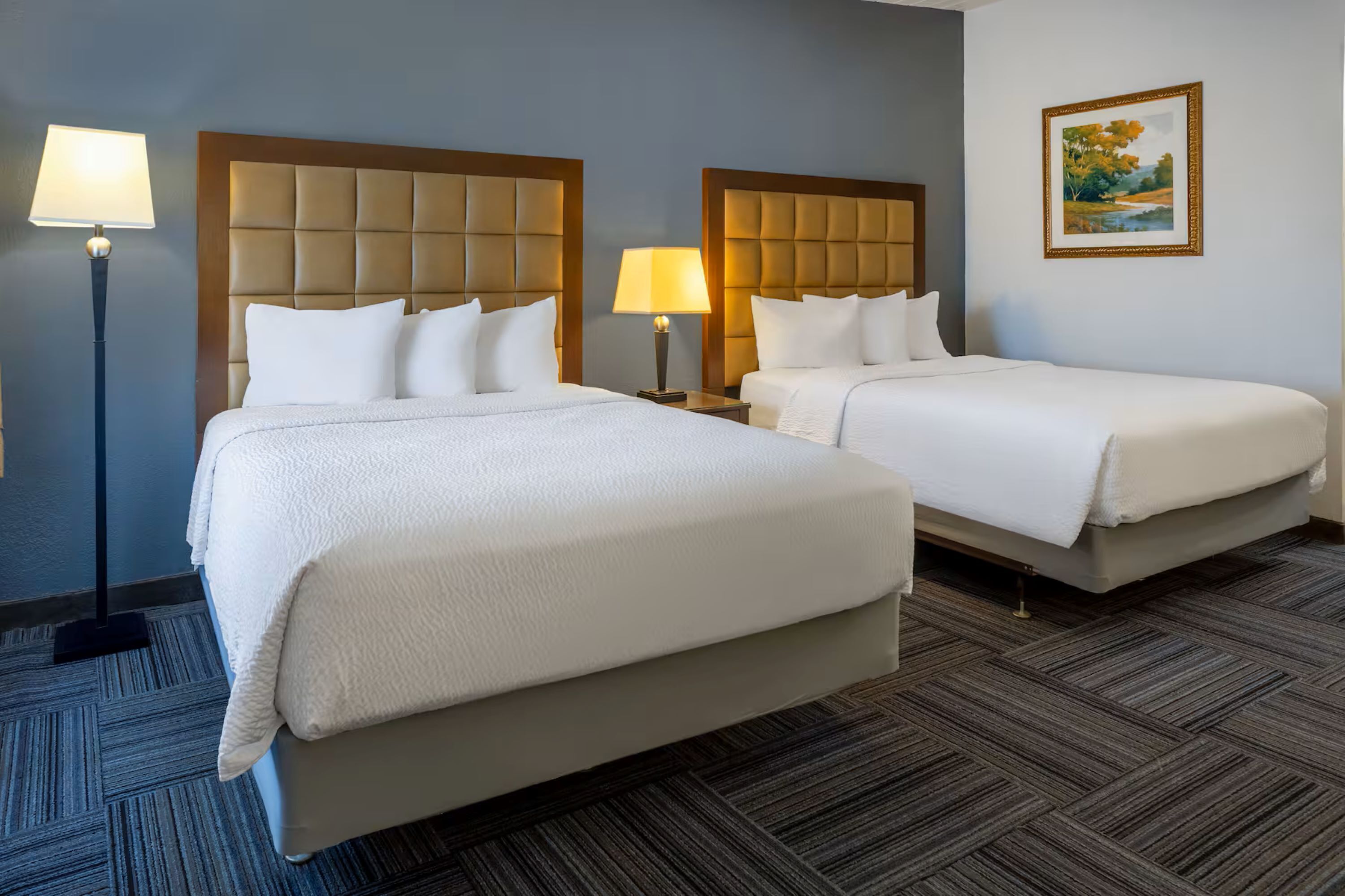 Quarto de hotel com duas camas, mesa de cabeceira com abajur, um quadro na parede e piso coberto por carpete.