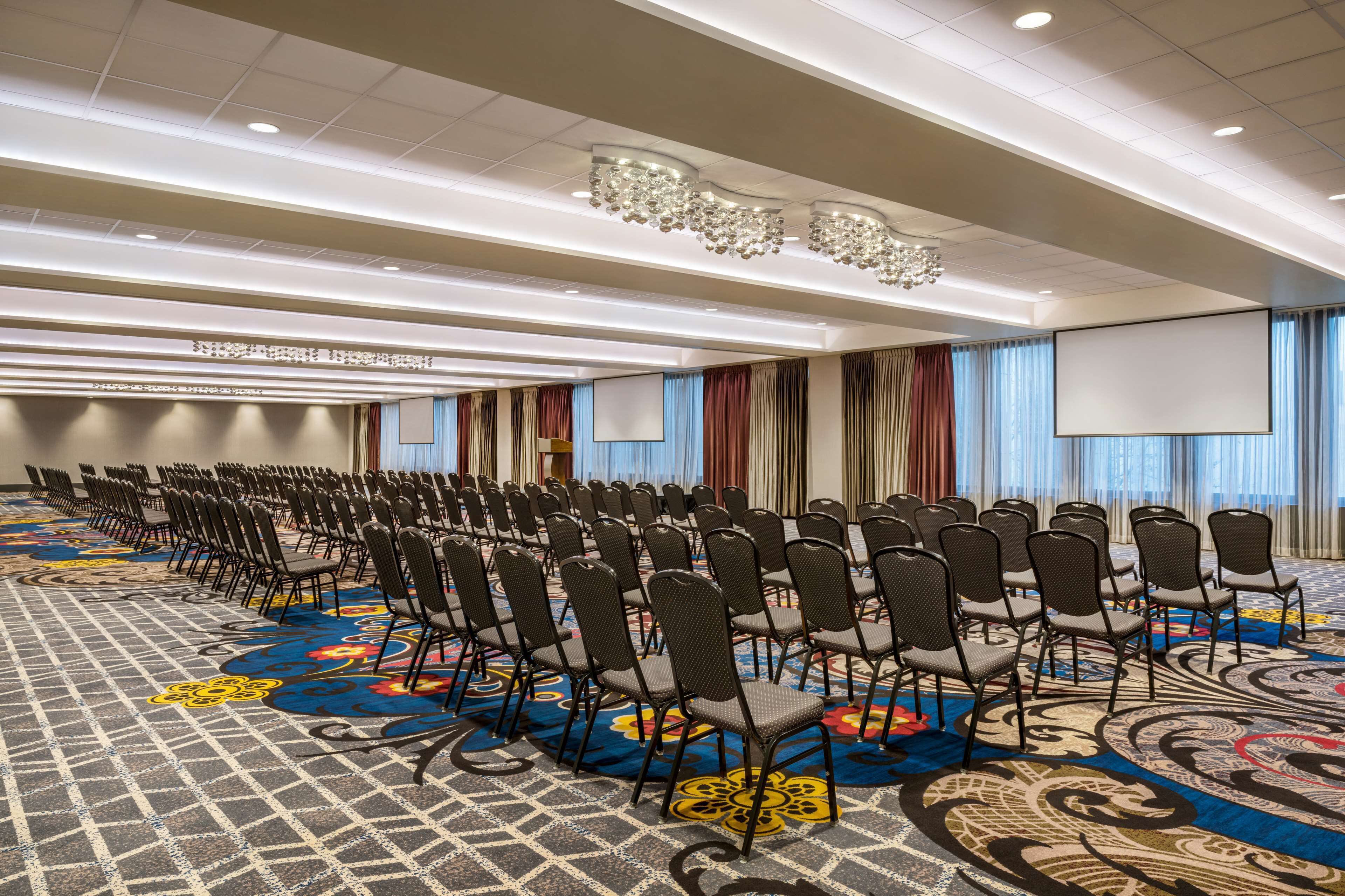 Un salón de conferencias con filas de sillas, una alfombra grande con estampado floral y una pantalla para proyector.