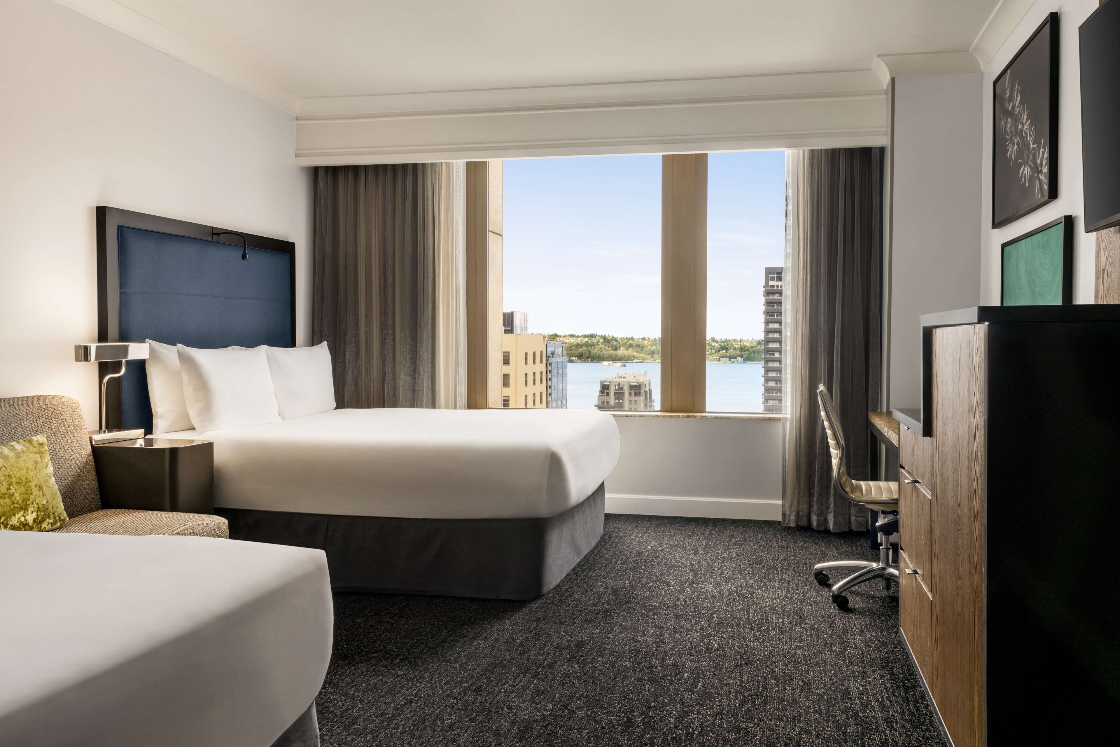 A hotel room with a large bed, a smaller bed, a desk, a chair, and a view of a body of water and buildings.