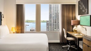 Une chambre d’hôtel équipée d’un lit, d’un bureau, d’une chaise, d’une lampe et d’une grande fenêtre donnant sur un immeuble et sur l’eau.