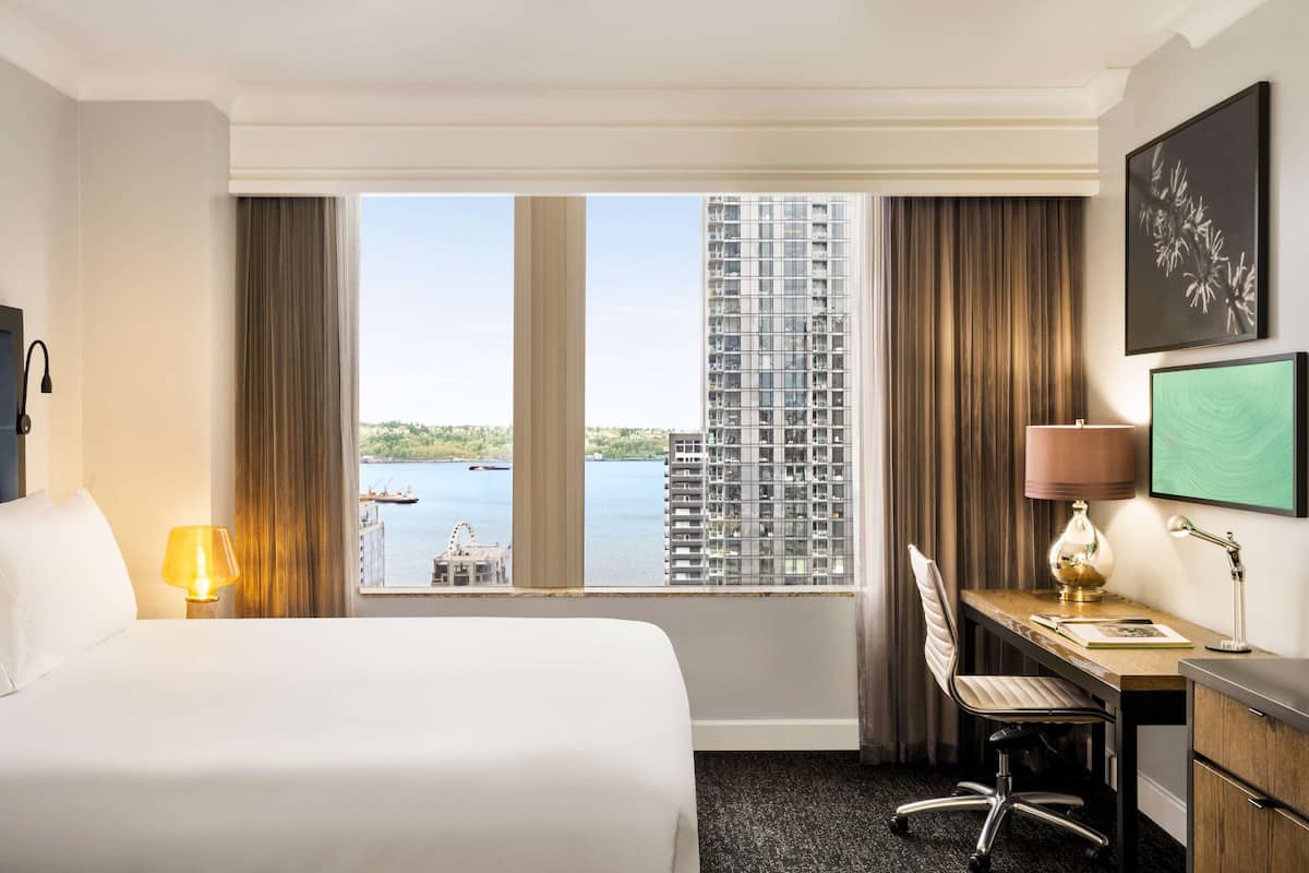 A hotel room with a bed, desk, chair, lamp, and a large window with a view of a building and water.