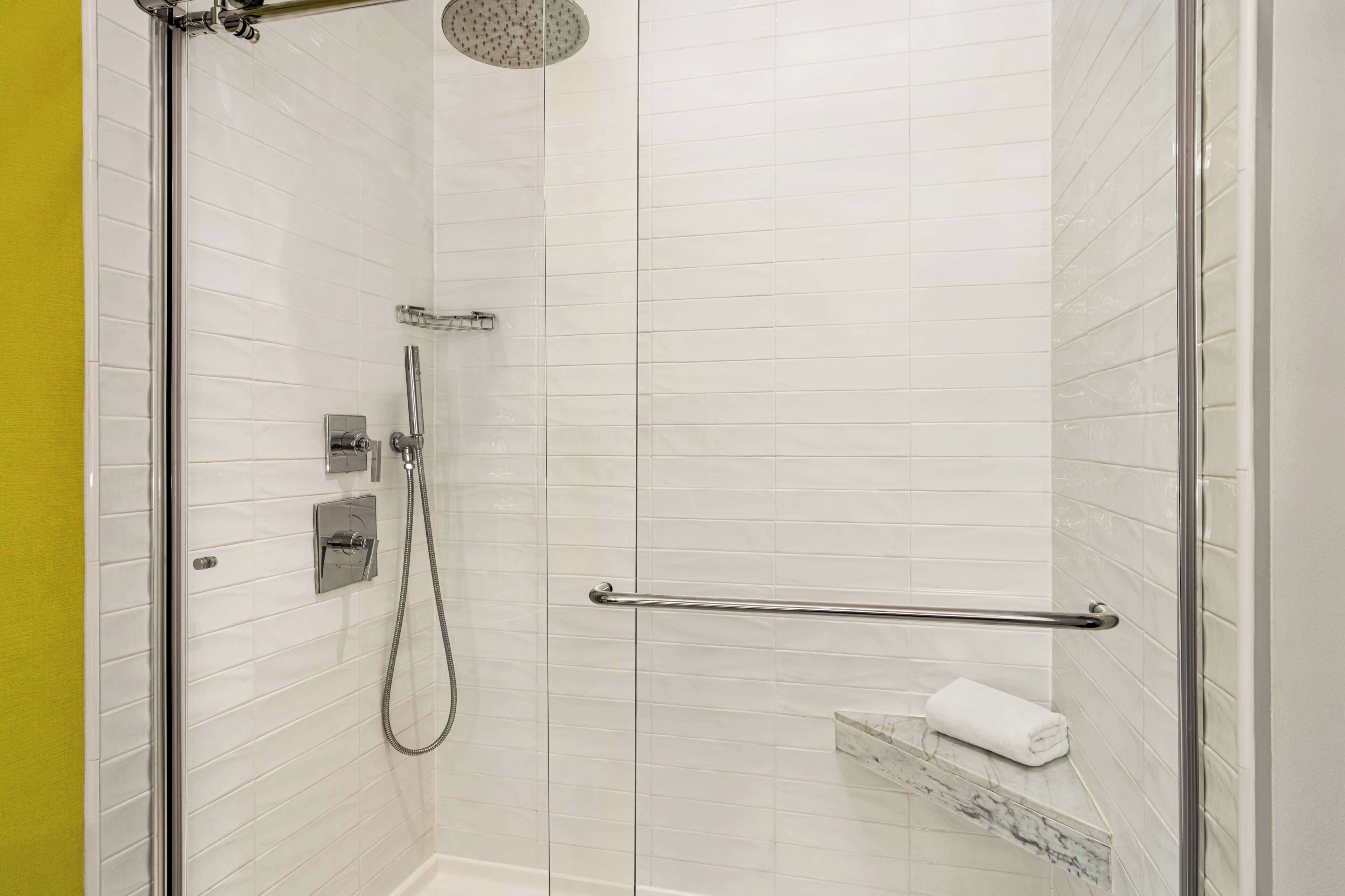 A modern shower with white tiles, a glass door, a handheld showerhead, a towel rack, and a marble accent on the floor.