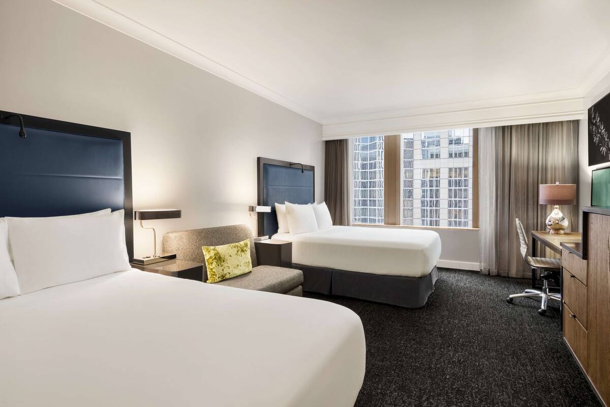 A hotel room with two beds, a desk, a chair, a lamp, and a large window with a city view.