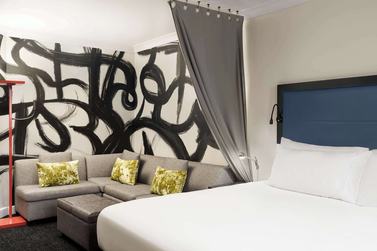 A modern hotel room with a sectional sofa, a large bed, and a wall with abstract black and white art.