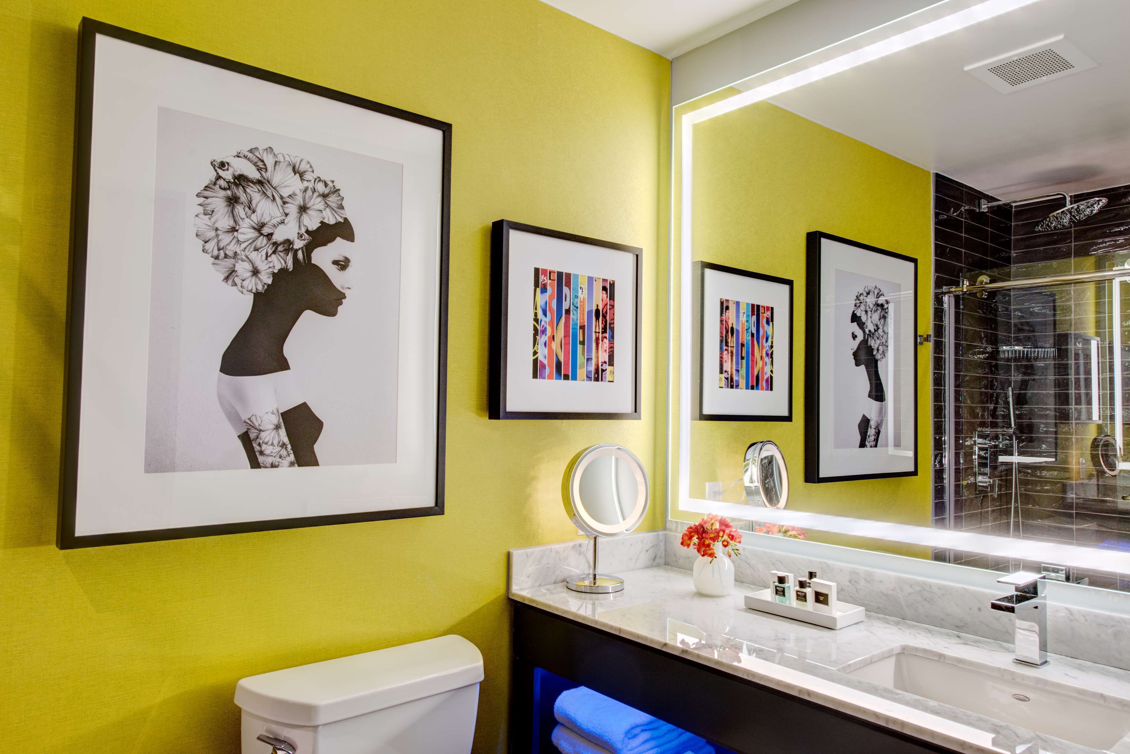 A modern bathroom with a yellow wall, a large mirror, framed artwork, a toilet, and a sink with a marble countertop.