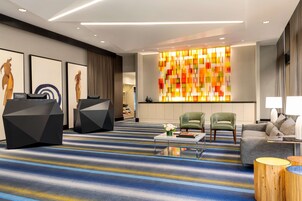 A modern hotel lobby with a striped carpet, two green armchairs, a grey sofa, a coffee table, and a colorful abstract wall.