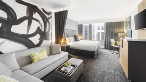 A modern hotel room with a large abstract painting, a sofa, a bed, a desk, and a TV.