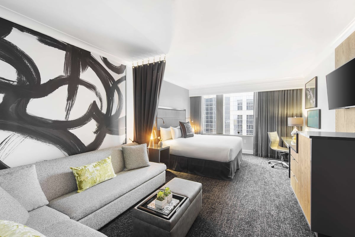 A modern hotel room with a large abstract painting, a sofa, a bed, a desk, and a TV.