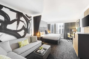 A modern hotel room with a large abstract painting, a sofa, a bed, a desk, and a TV.