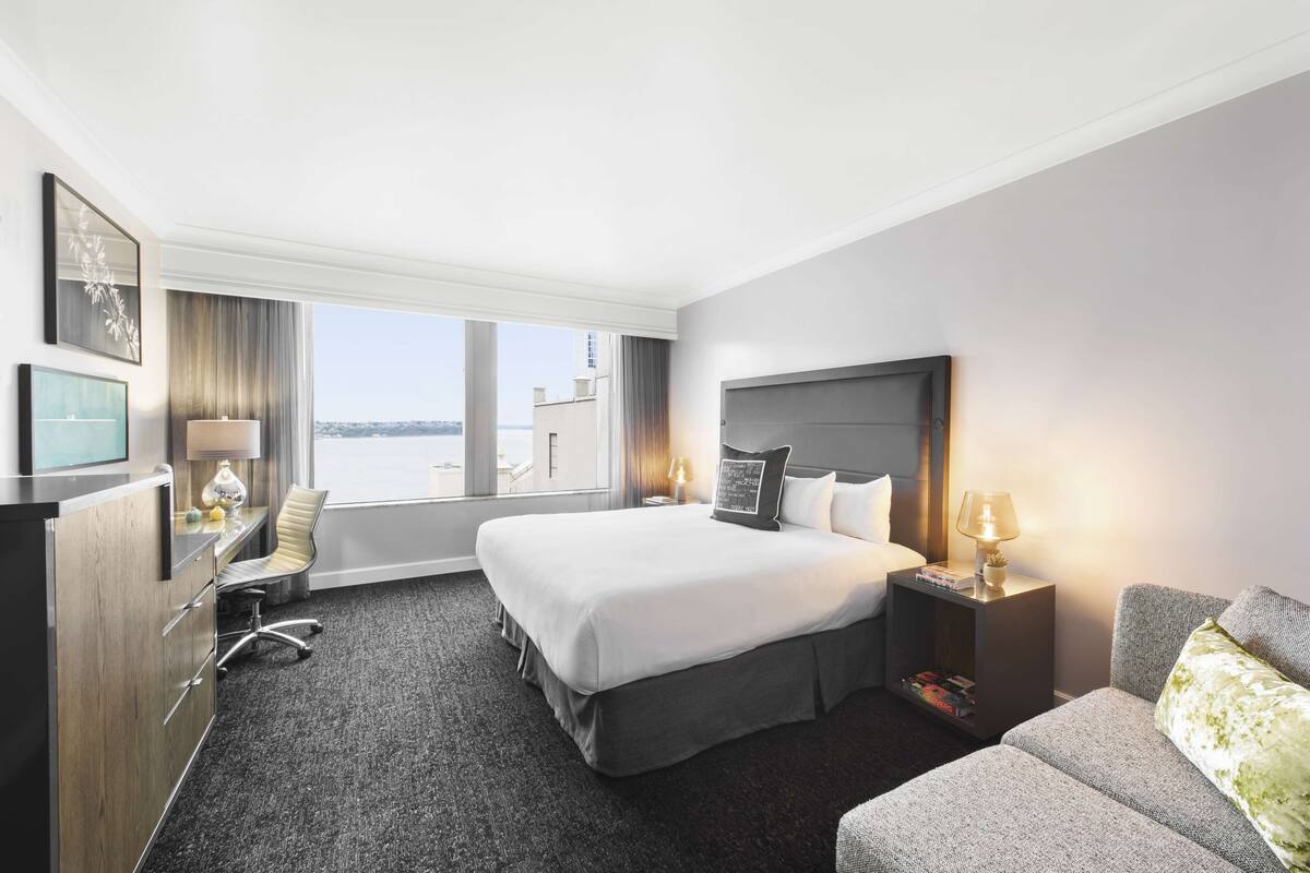 A hotel room with a large bed, a desk, a chair, a TV, and a view of the city.