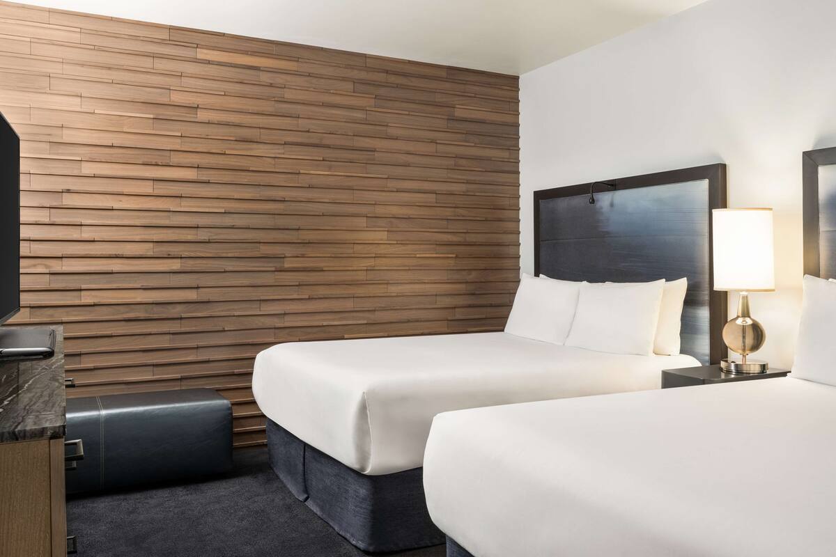 A hotel room with two beds, a wooden accent wall, a flat-screen TV, a bedside table with a lamp, and a small seating area.