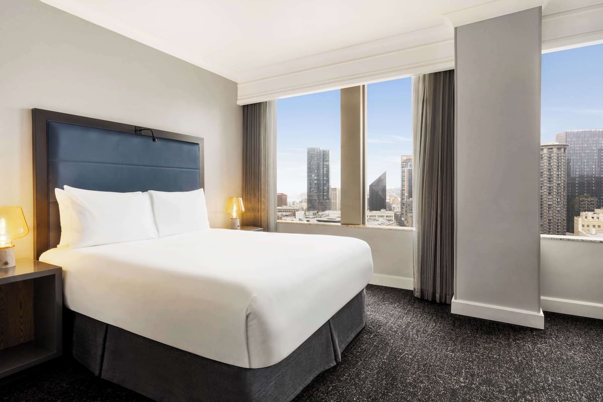 A hotel room with a large bed, a nightstand, a lamp, and a view of the cityscape through the window.