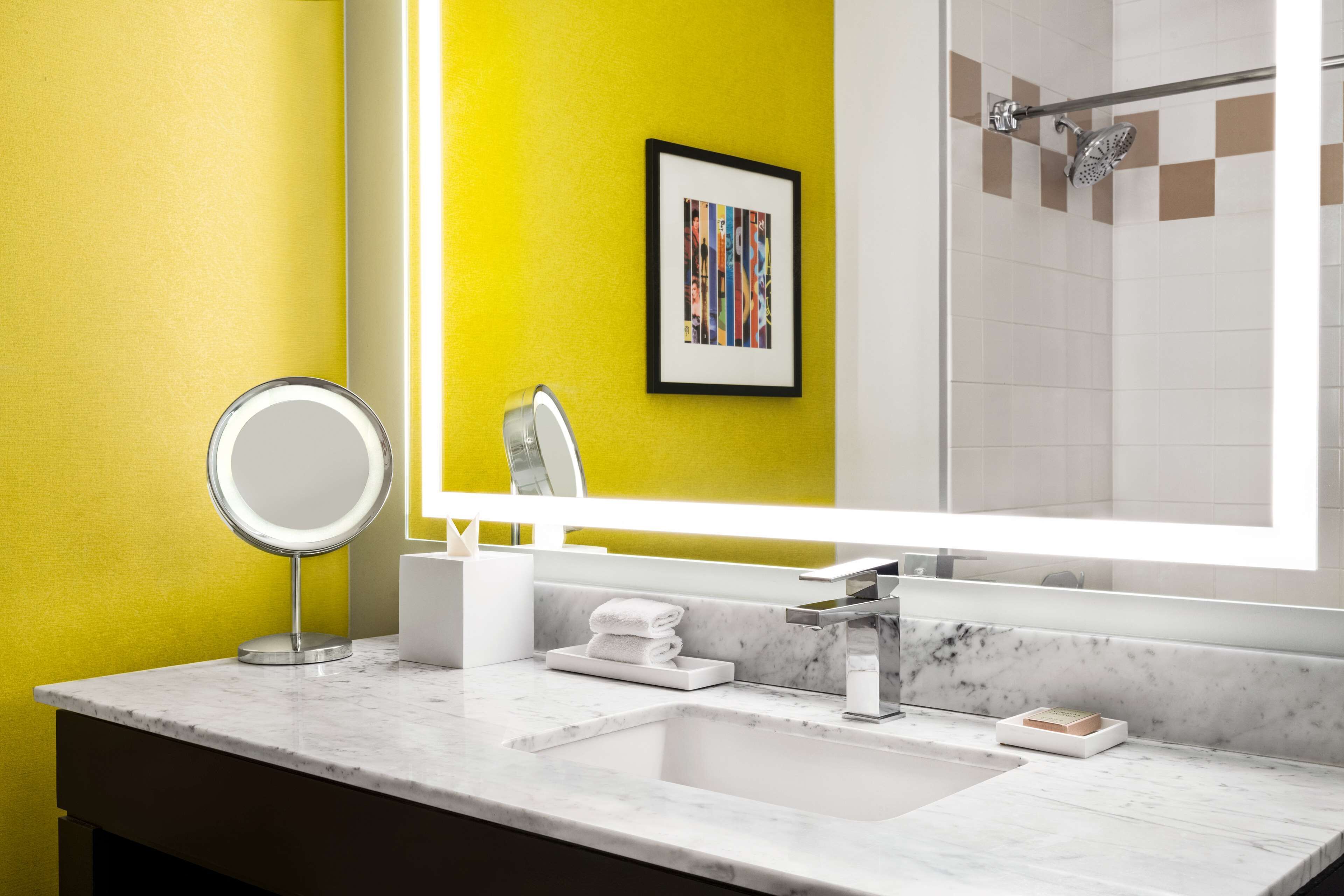 Un baño moderno con una pared de acento amarilla, un espejo grande, un lavabo con encimera de mármol y una zona de ducha.
