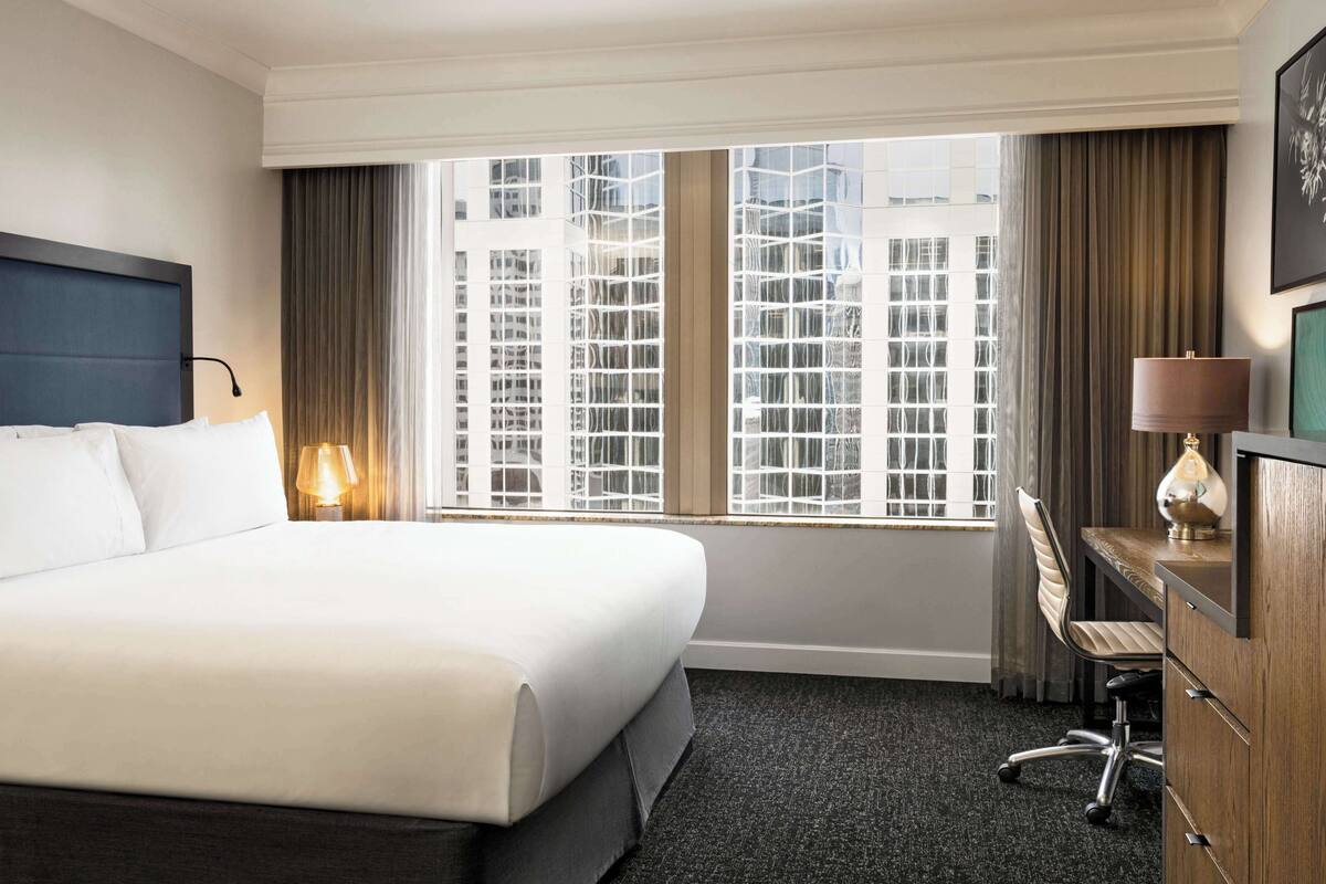 A hotel room with a large bed, a desk with a chair, a lamp, and a view of buildings through the window.
