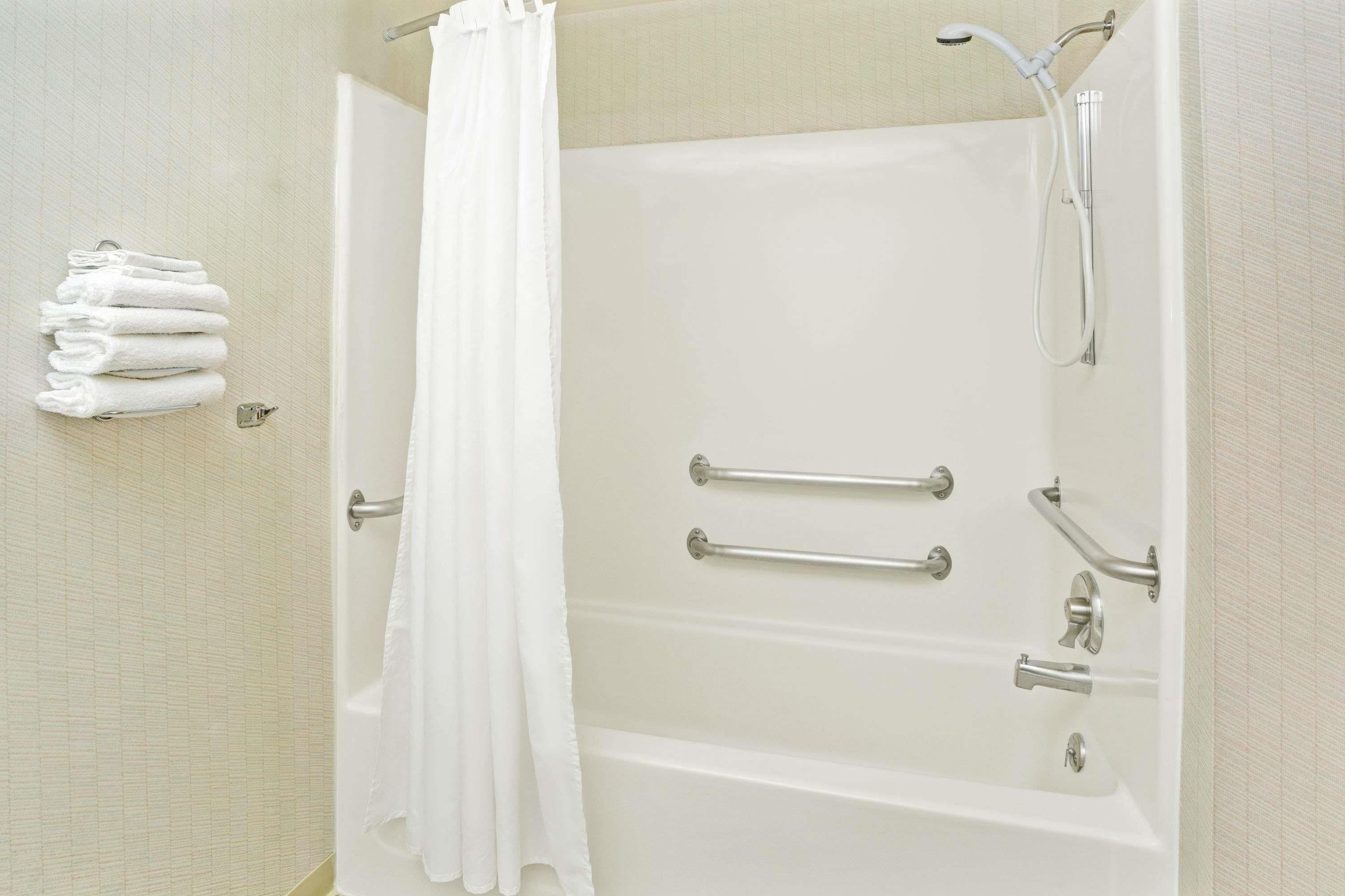A bathroom with a bathtub, grab bars, a showerhead, and a stack of white towels.