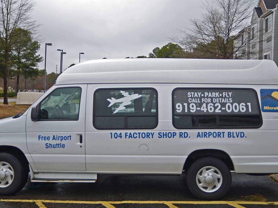 Una camioneta blanca con la leyenda "Free Airport Shuttle" y "104 FACTORY SHOP RD. AIRPORT BLVD." en la carrocería.