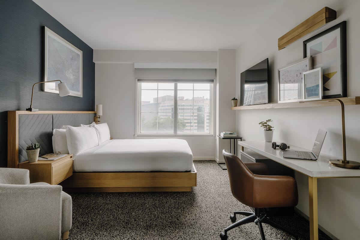 A modern hotel room with a bed, desk, chair, and a large window offering a city view.