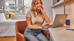 Eine Person sitzt an einem Schreibtisch mit einem Laptop in einem modernen Zimmer mit Bett, Fenster und Lampe.