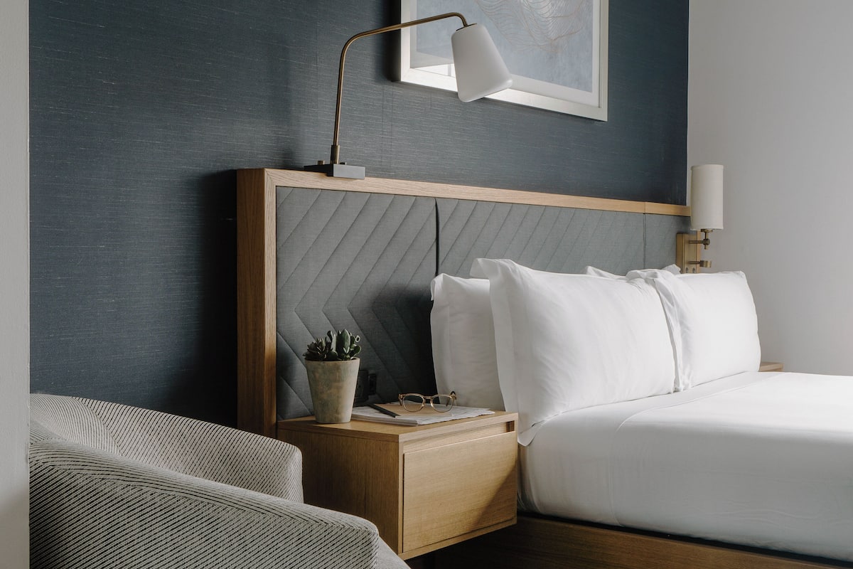 A neatly made bed with white linens, a wooden headboard, a bedside table with a lamp, a potted plant, and a framed picture on the wall.