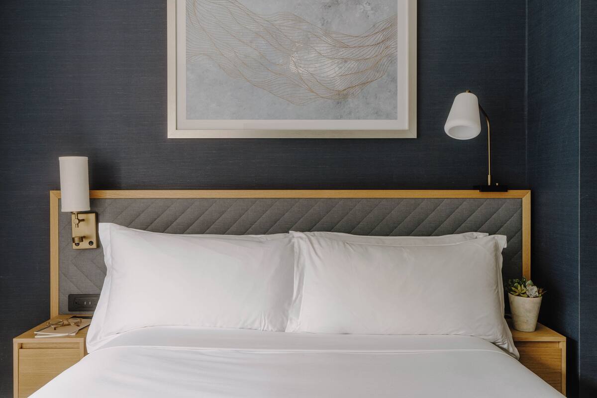 A neatly made bed with white linens, a headboard with a textured pattern, a bedside lamp, a framed artwork above the bed, and a small potted plant on the nightstand.