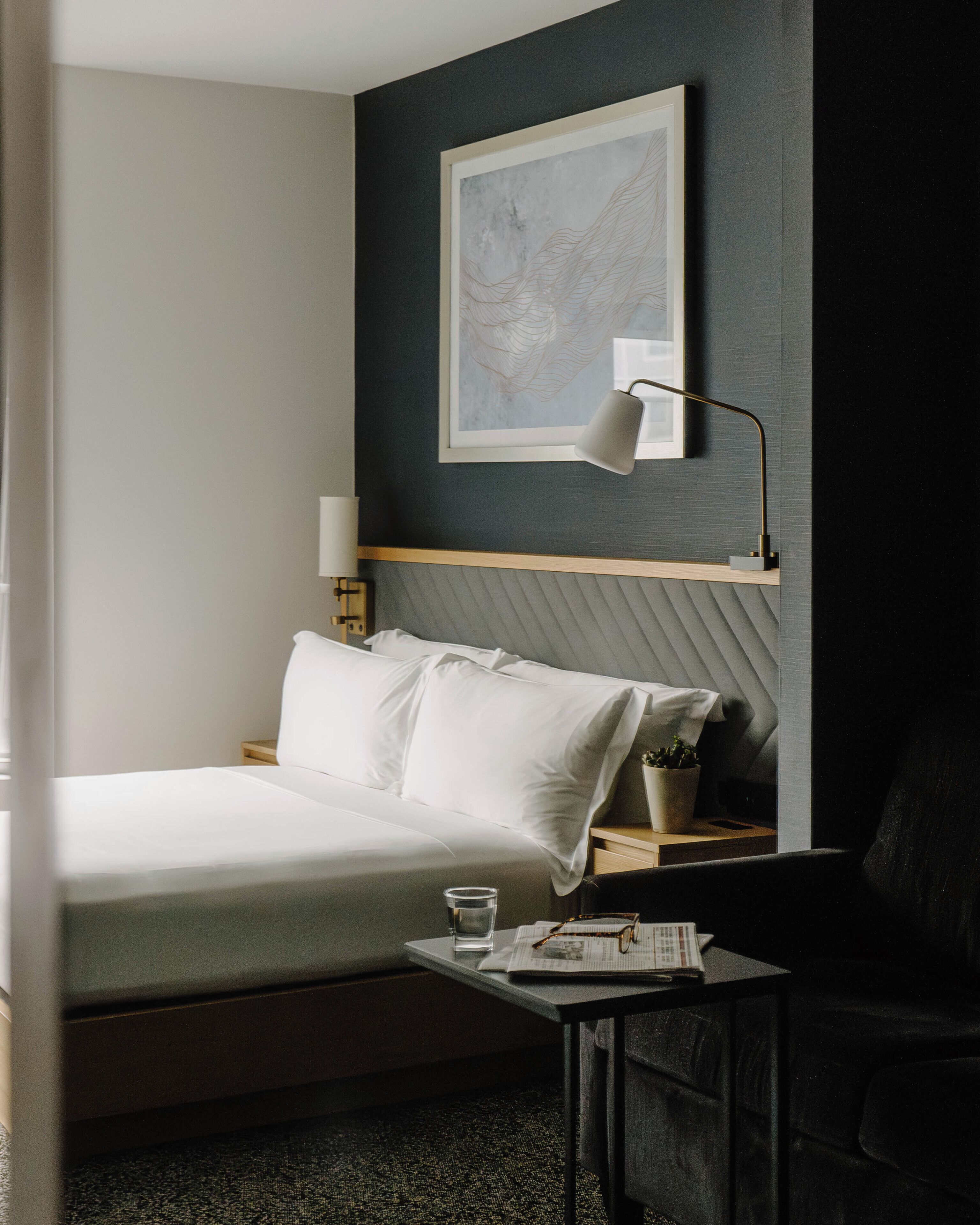 Siististi järjestetty hotellihuone, jossa on sänky, yöpöytä, lamppu ja seinällä oleva kehystetty taideteos.