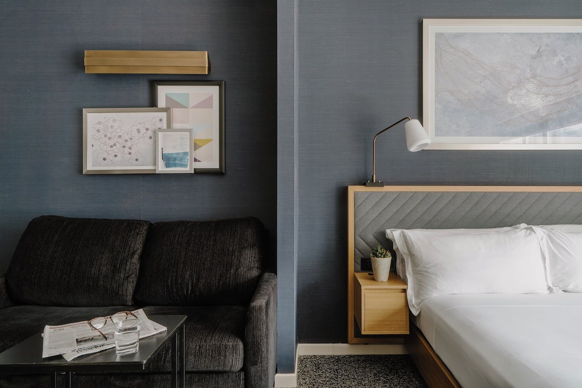 A modern hotel room with a dark grey wall, a grey sofa, a wooden bedside table with a lamp, a bed with white linens, and framed artwork above the sofa.