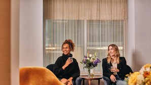 Zwei Personen sitzen auf einem Sofa in einem Zimmer mit einem großen Fenster, einem kleinen Tisch mit Blumenvase und einem gemusterten Teppich.
