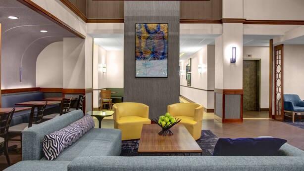 A hotel lobby with seating areas, a central table with a fruit bowl, and a large abstract painting on the wall.