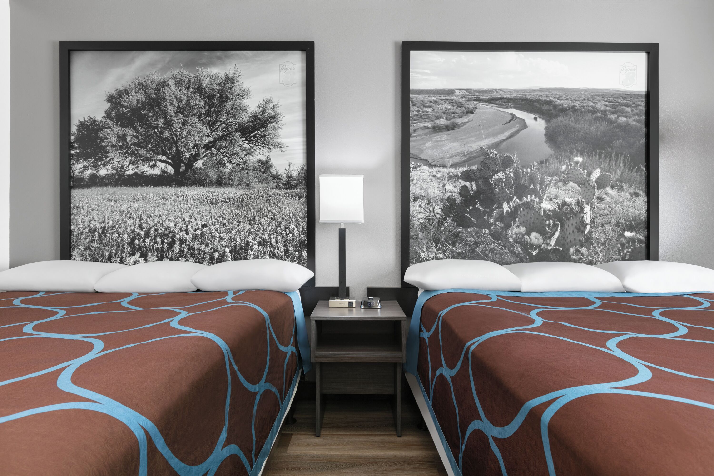 Habitación de hotel con dos camas, una mesita de noche, una lámpara y dos fotografías en blanco y negro de paisajes enmarcadas en la pared.