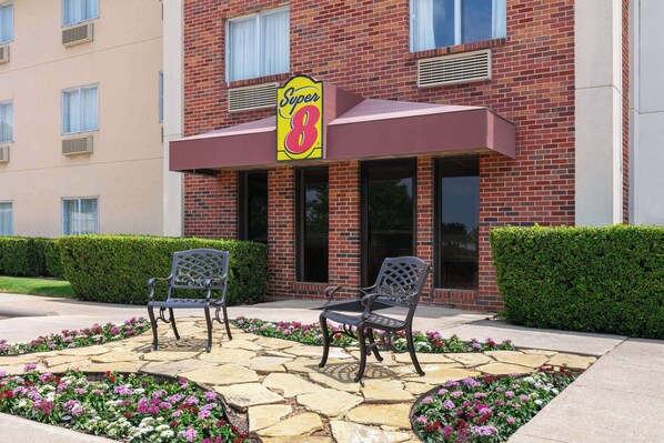 A Super 8 hotel exterior with a brick facade, two outdoor seating areas with chairs, and a flower bed in front. - Super 8 by Wyndham Grapevine/DFW Airport Northwest (Grapevine)