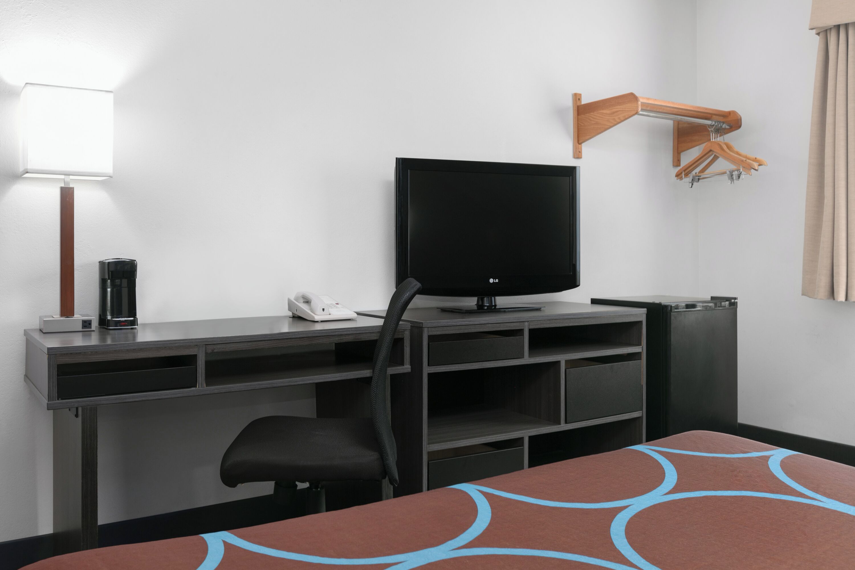 A hotel room with a flat-screen TV on a stand, a desk with a telephone, a chair, and a small table with a patterned table runner.