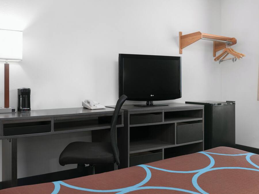 A hotel room with a flat-screen TV on a stand, a desk with a telephone, a chair, and a small table with a patterned table runner.