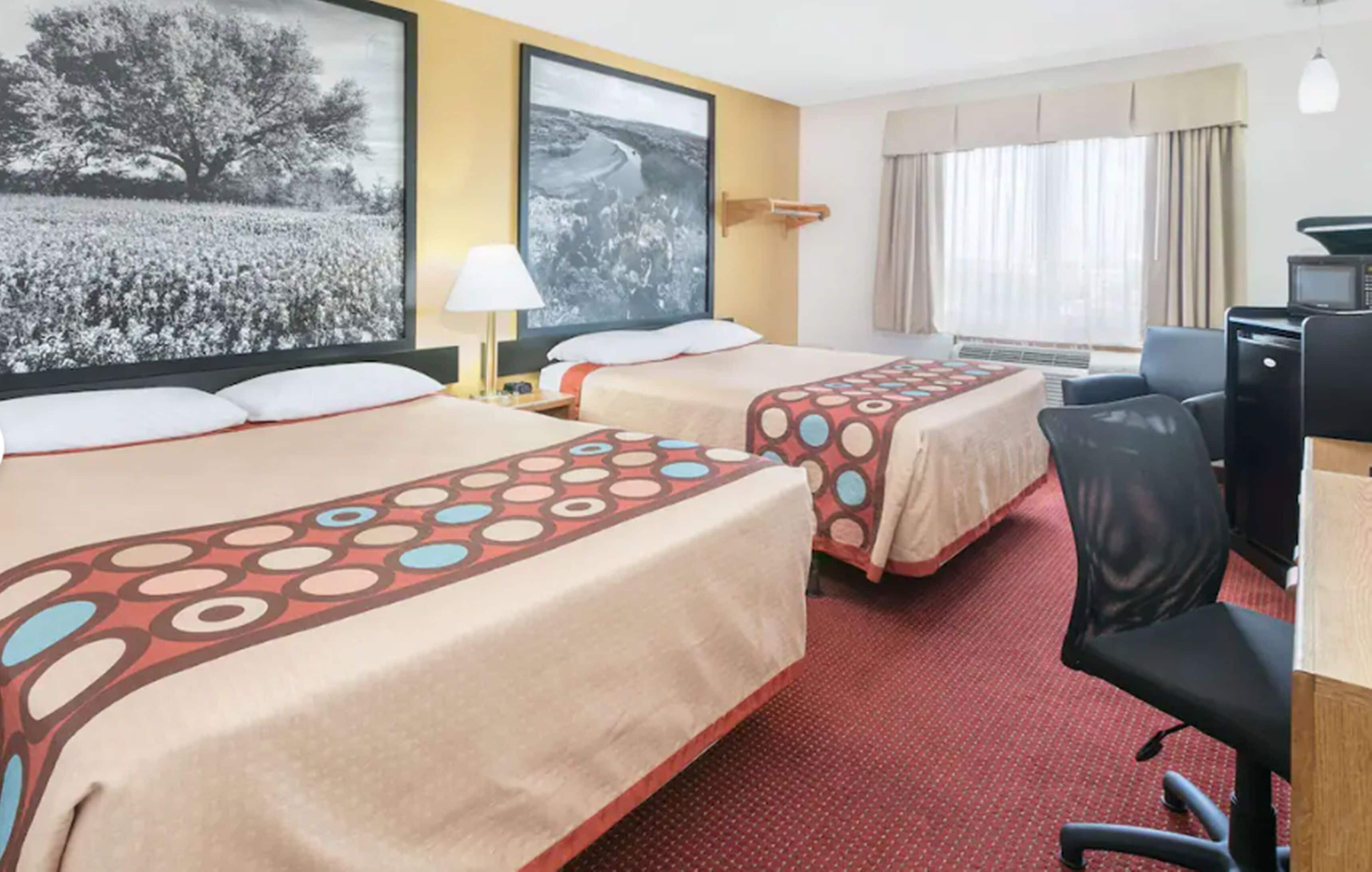 Habitación de hotel con dos camas, un escritorio, una silla, televisión y un ventanal con cortinas.