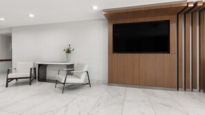Una moderna zona de recepción con suelo de mármol, dos sillas blancas, una pequeña mesa con un florero y un televisor de pantalla plana grande montado sobre un panel de madera.