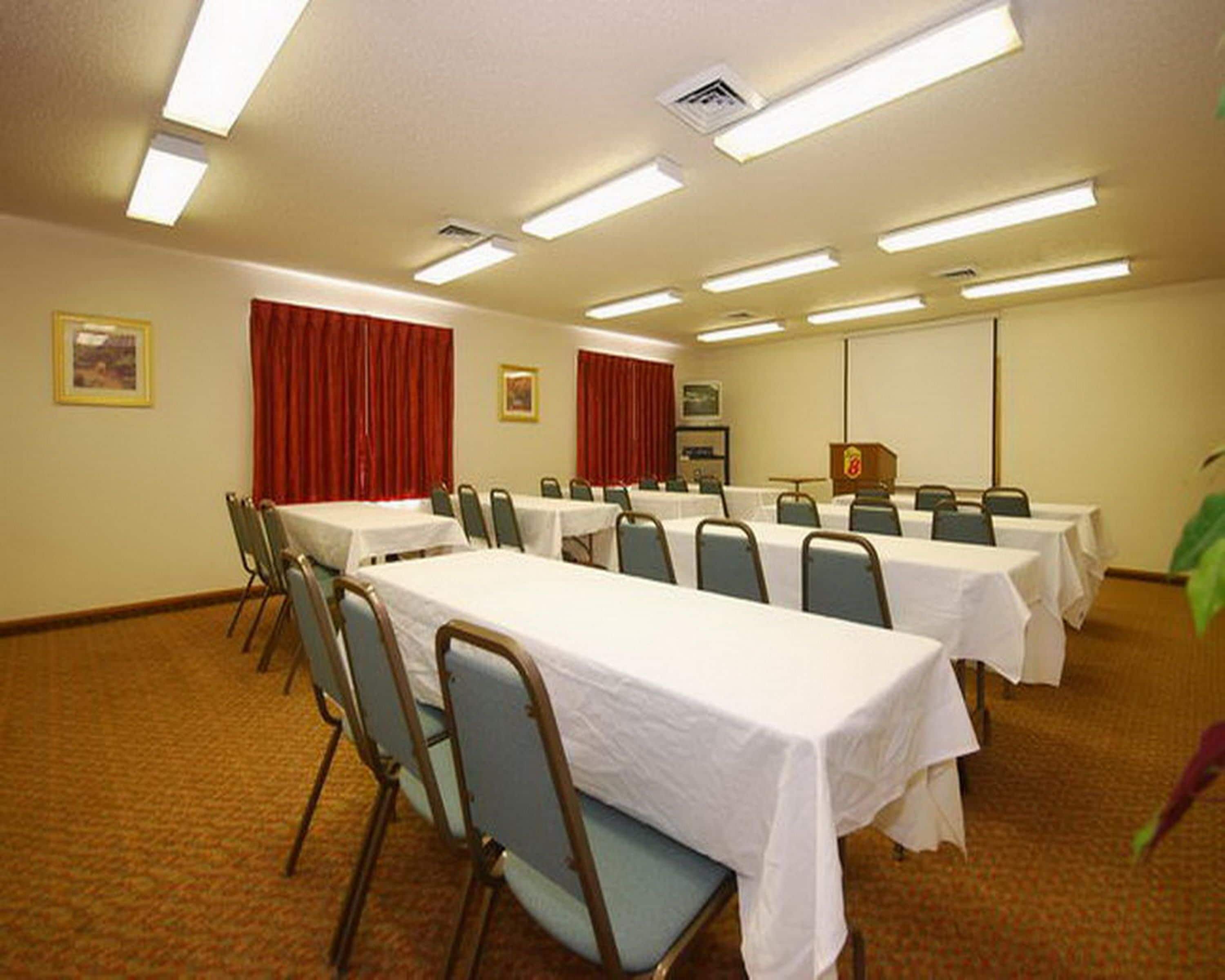 Un salón de conferencias con varias mesas y sillas, equipado con luces en el techo y una pantalla de proyector.