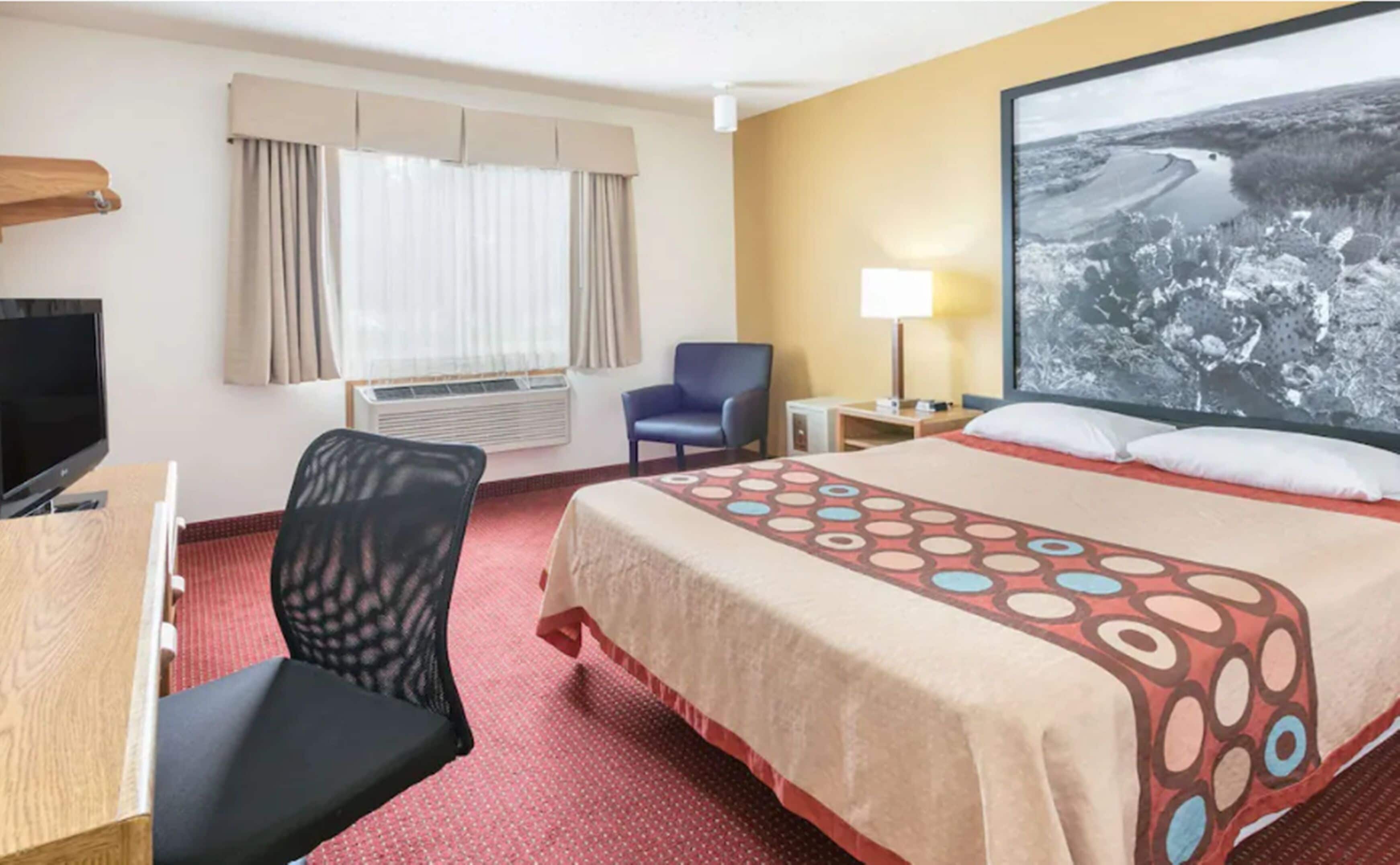 A hotel room with a bed, a desk, a chair, a TV, a window with curtains, an air conditioner, a lamp, and a large picture on the wall.