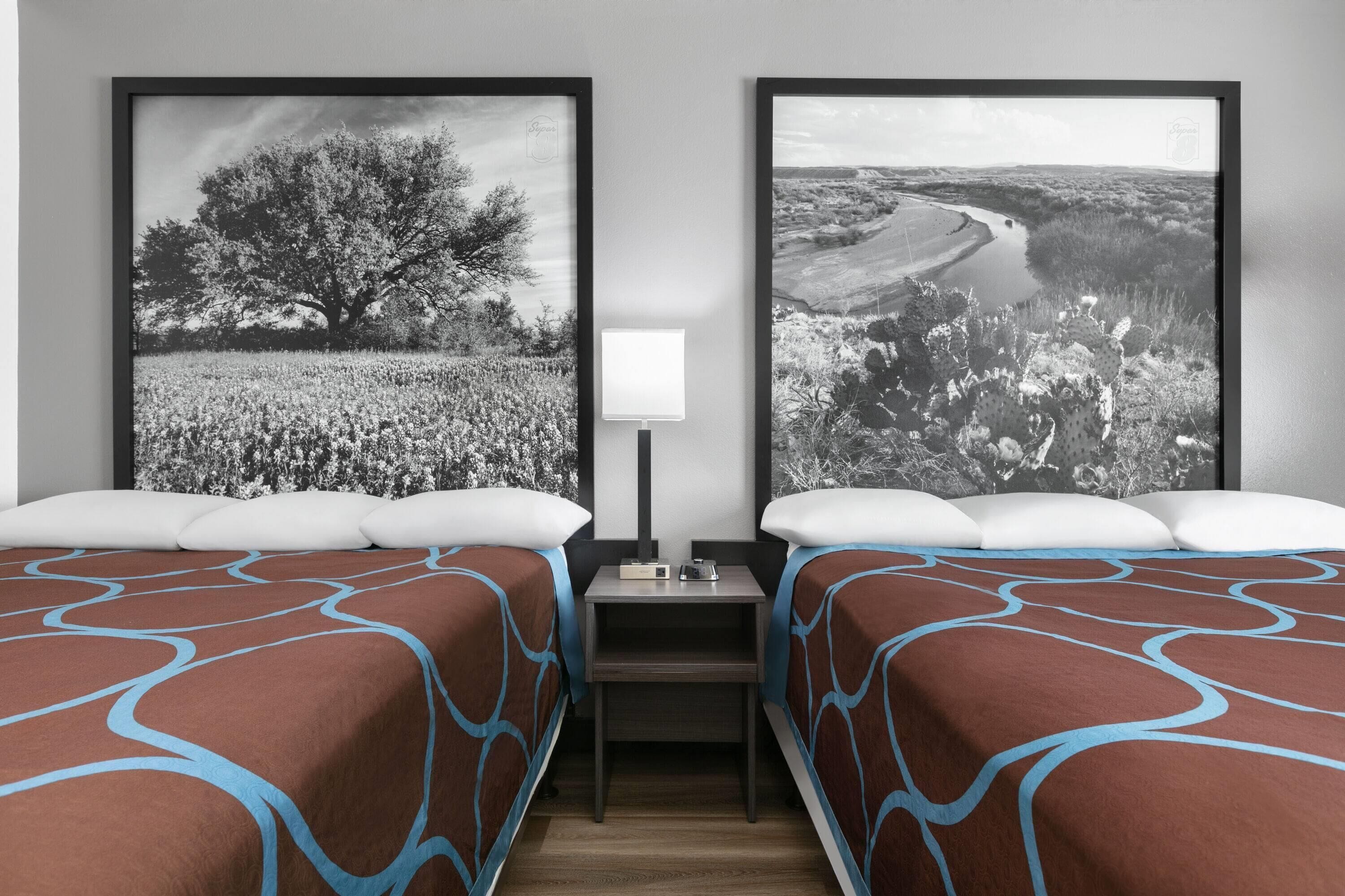A hotel room with two beds, a nightstand, a lamp, and two framed black and white landscape photographs on the wall.