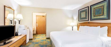 A hotel room with a bed, a television on a wooden dresser, a desk with a lamp, a microwave, and a door leading to another room.