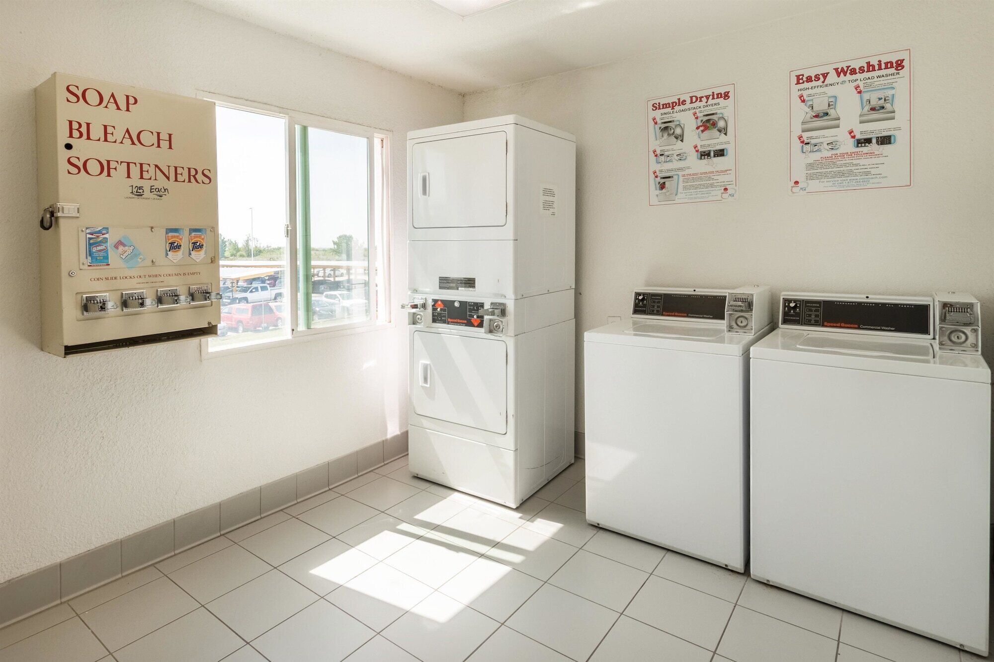 A laundry room with two washers and dryers, a coin-operated soap and bleach softener dispenser, and a window with a view of a parking lot.