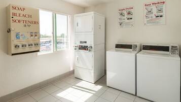 A laundry room with two washers and dryers, a coin-operated soap and bleach softener dispenser, and a window with a view of a parking lot.