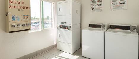 A laundry room with two washers and dryers, a coin-operated soap and bleach softener dispenser, and a window with a view of a parking lot.